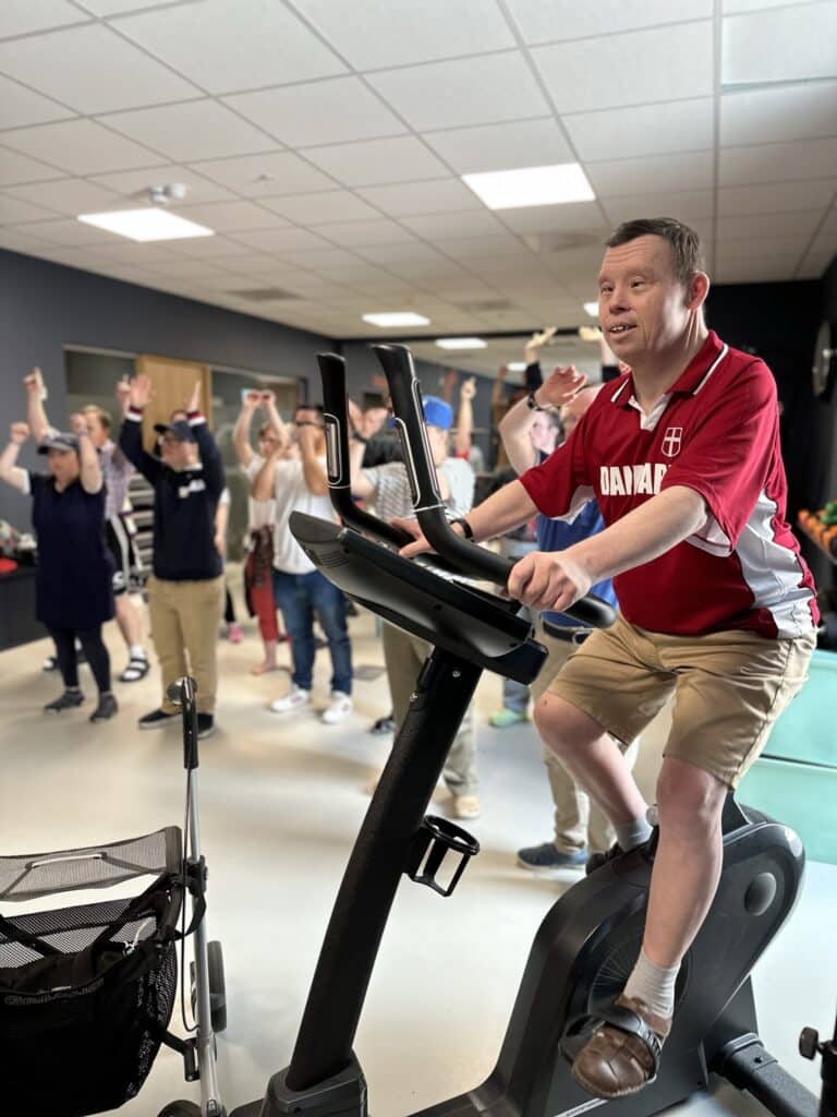 En mann som sykler på en ergometersykkel i et rom sammen med andre